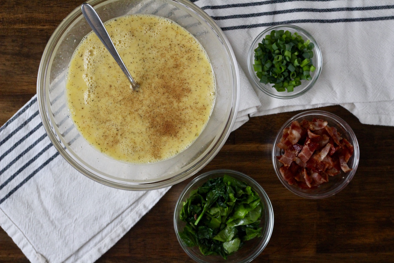 A large bowl with the eggs mixed together, a bowl with the green onions, and bacon.