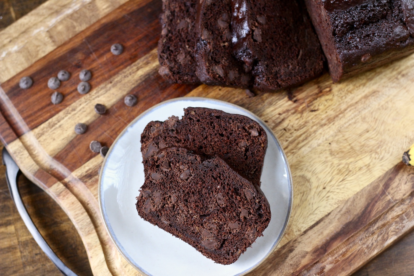 Two slices of chocolate banana bread on a small plate with some sliced in the back on a wooden cutting board.