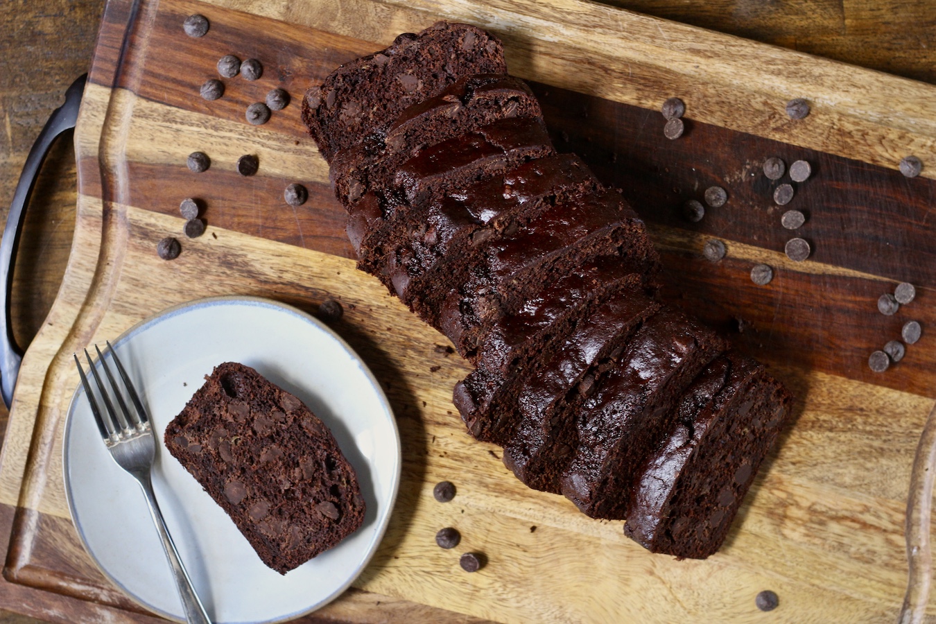 A loaf of chocolate banana bread sliced on a wooden cutting board with a small plate with a slice of bread on a plate to the left.