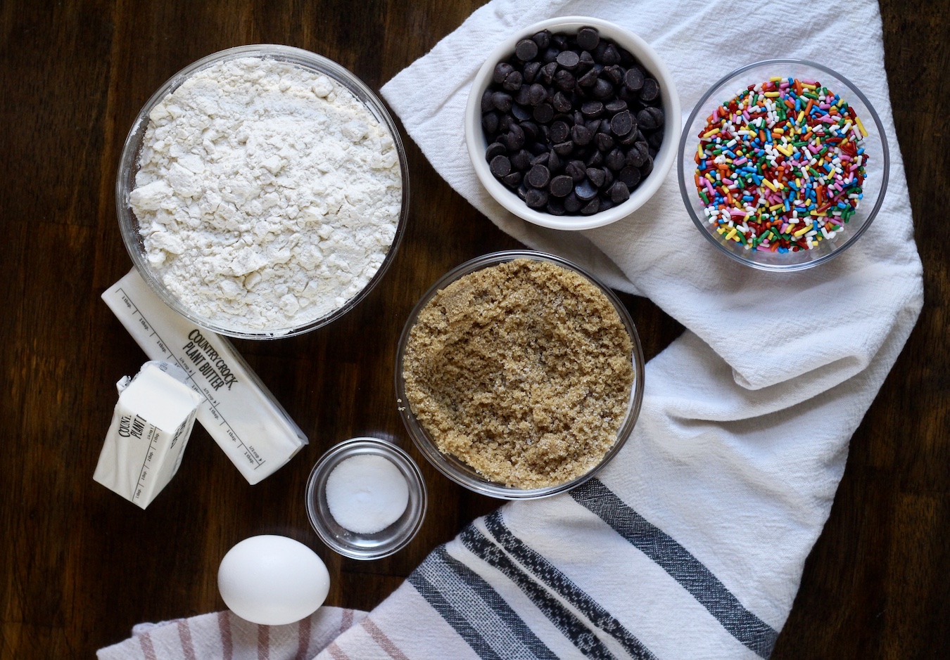 Ingredients in small bowls for Funfetti Cookie Cake on a wooden counter with a white and blue dish towel.