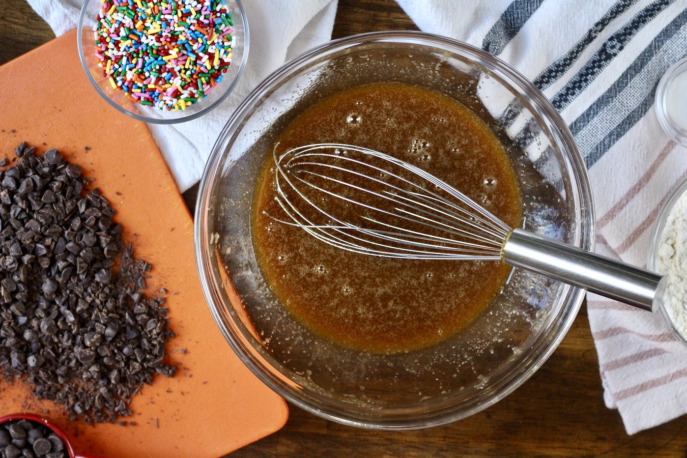 A glass bowl with melted butter mixed with sugar and a wire whisk. On the left is a pile of chopped chocolate and a bowl of sprinkles.