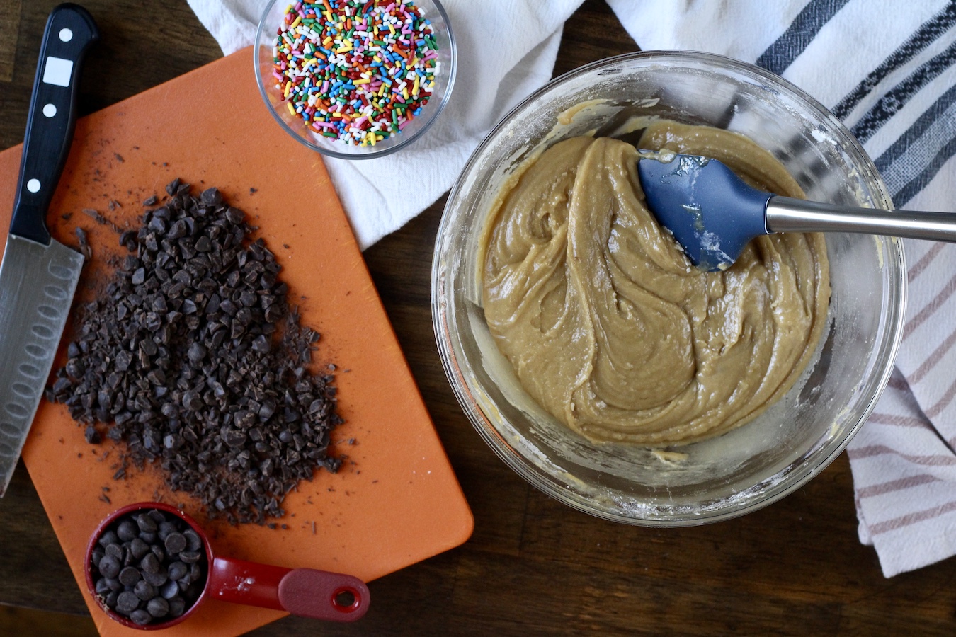 Cookie cake batter in a glass bowl with a spatula with a cutting board of chocolate to the left and a small bowl of sprinkles up top.