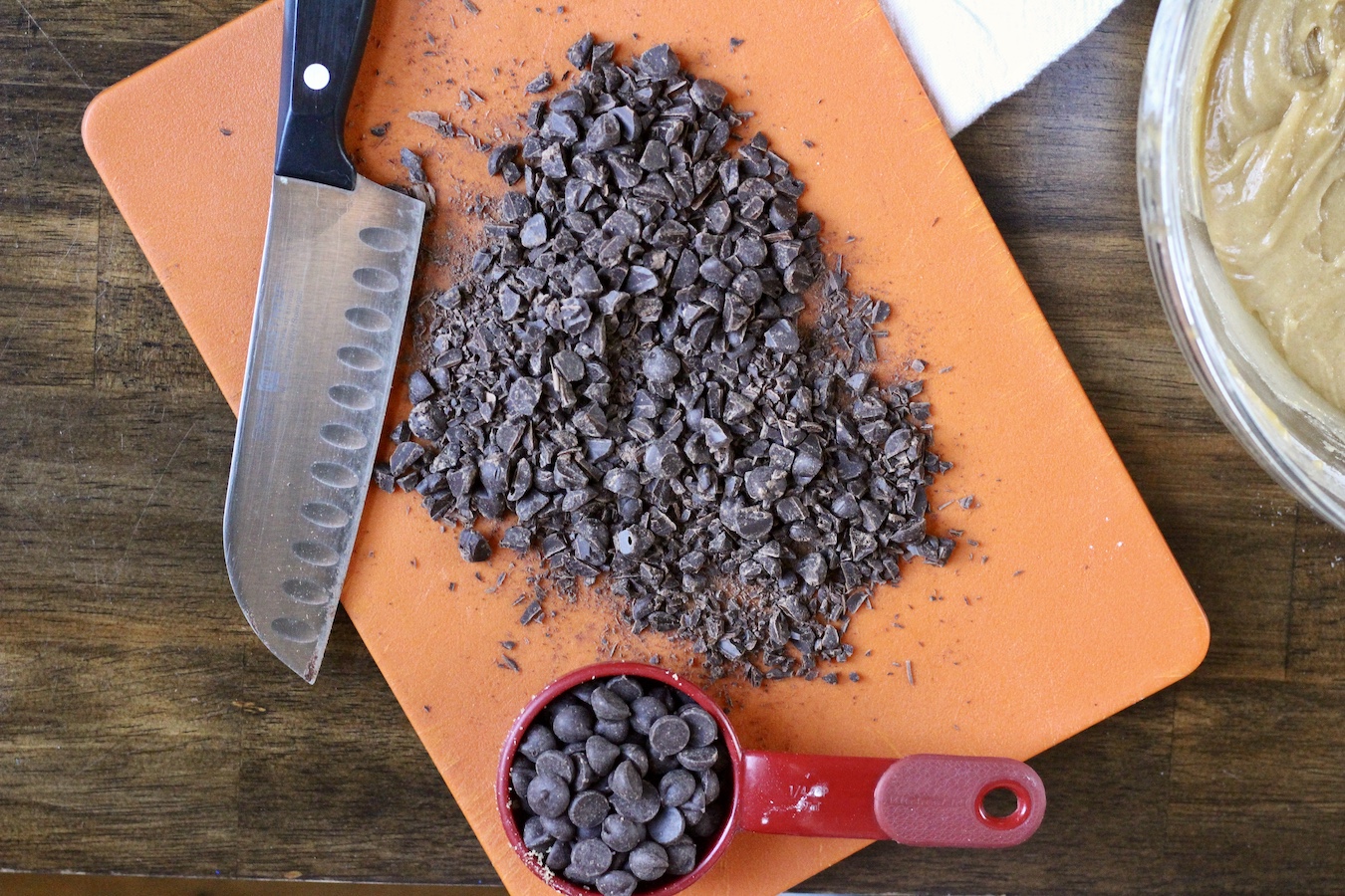 A wooden counter with a cutting board with chopped chocolate chips and a knife.