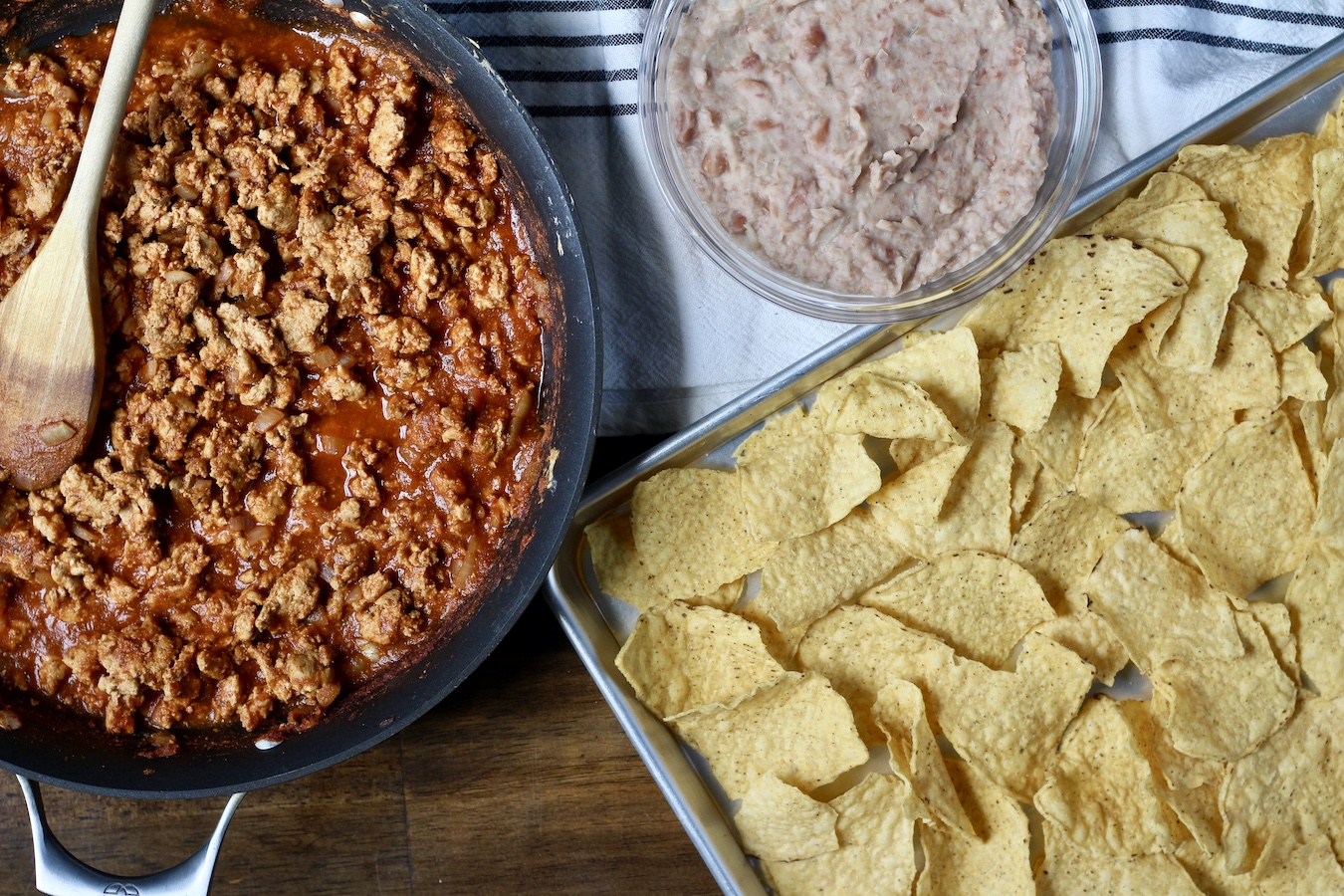 A sheet pan with tortilla chips on the right, a pan with taco meat on the left, and a bowl of refried beans at the top.