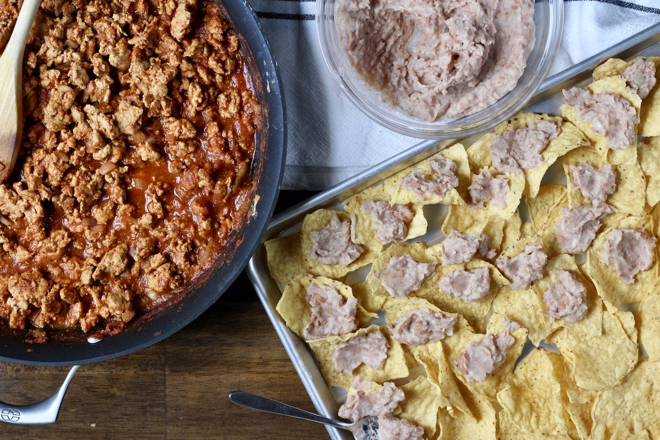 A sheet pan with tortilla chips topped with refried beans with a pan of taco meat.