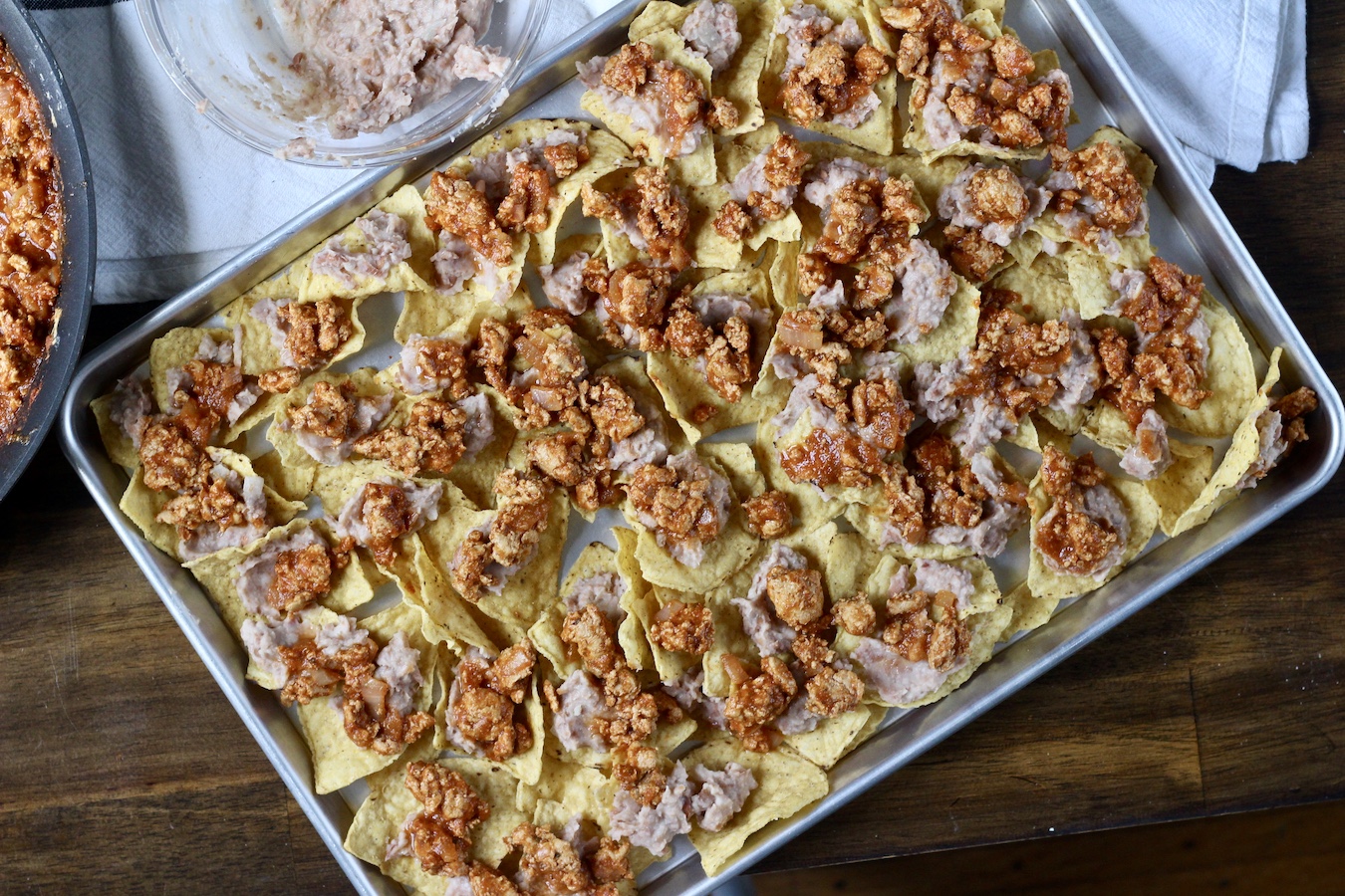 Nachos topped with refried beans and taco meat on a sheet pan.