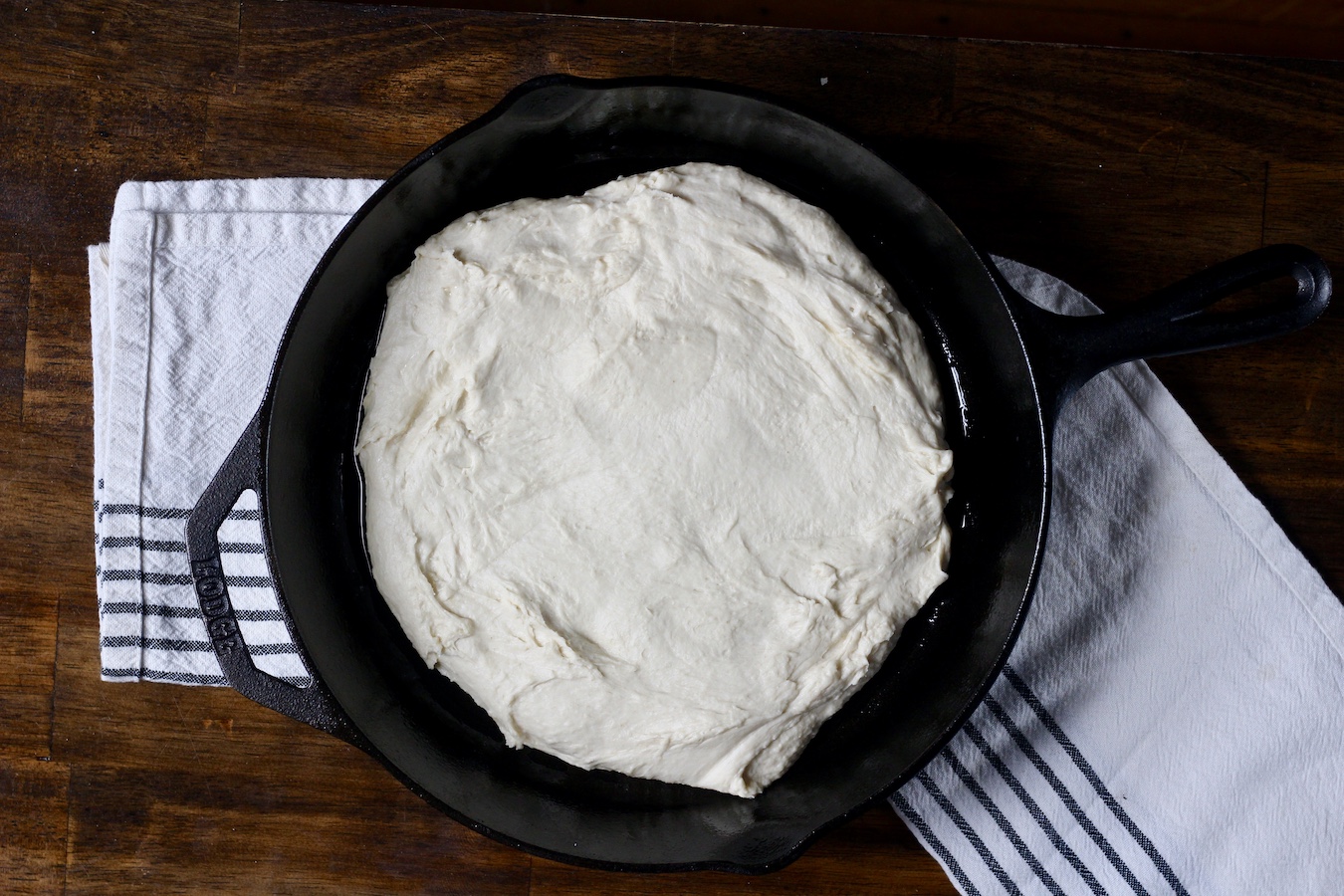 Focaccia dough in a cast iron skillet on a white a blue dish towel on a wooden table.