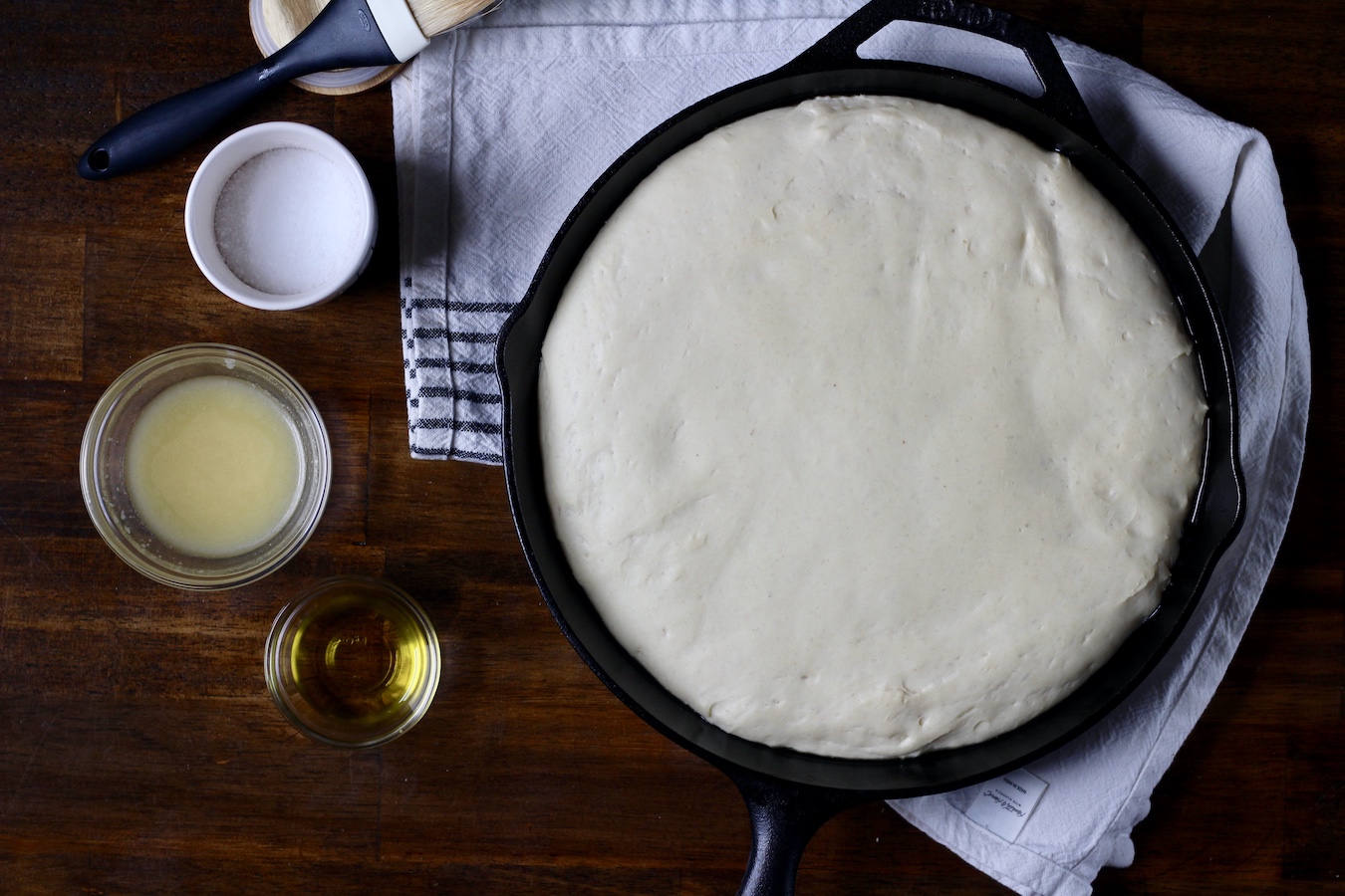 Focaccia dough in a cast iron skillet after rising for an hour with salt, vegan butter, and olive oil to the left.