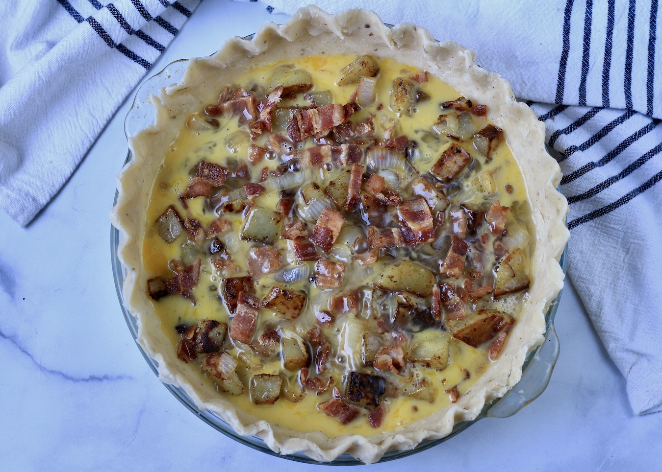 Uncooked bacon and potato quiche on a marble background.