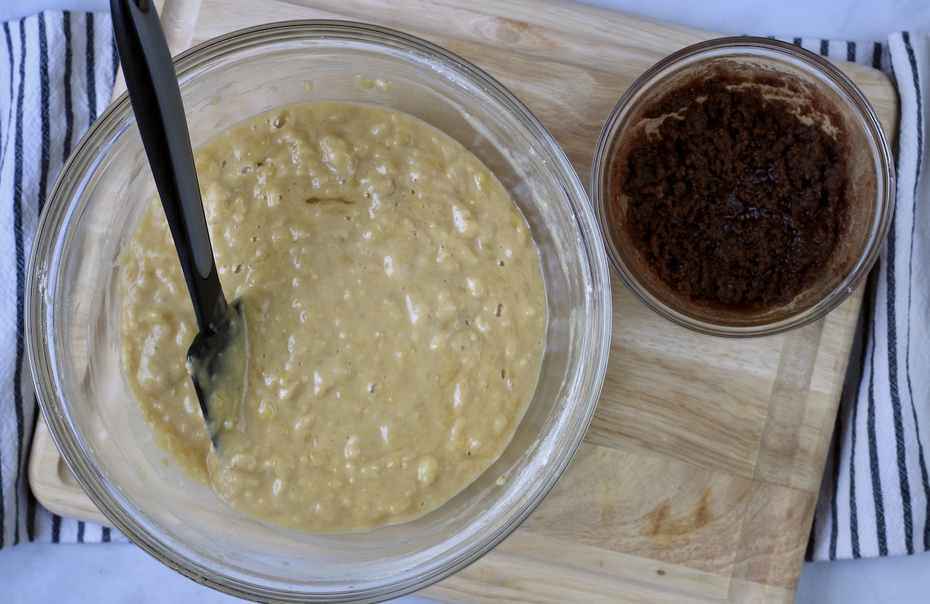 Banana bread batter in a large bowl with cinnamon filling in a small bowl on the right in a wooden counter.