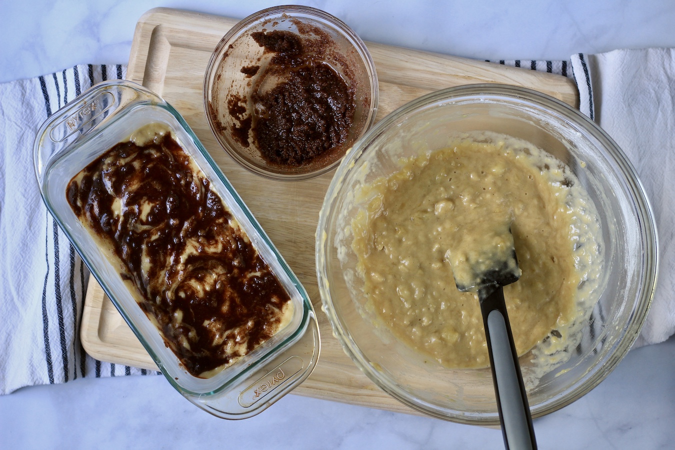 A bread pan with cinnamon swirl batter and two bowls with banana batter and cinnamon filling on a wooden counter.