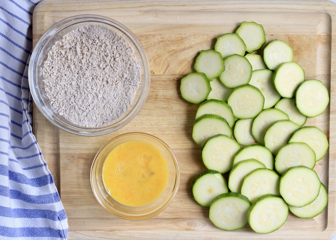 Sliced zucchini on a cutting board with a small bowl with whisked egg and another bowl with the breading mixture.