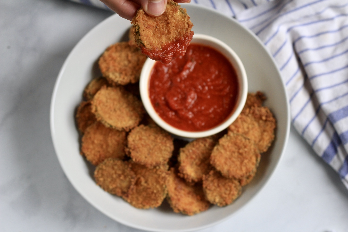 A white bowl with zucchini bites and a small bowl of marinara with a hand holding an oven baked zucchini bite that was dipped in marinara sauce.