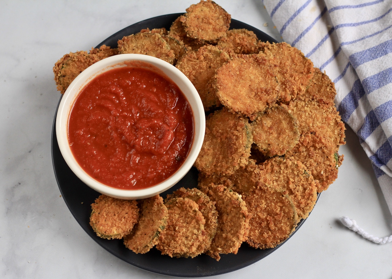 A blue plate with a pile of zucchini bites on the right and a small bowl of marinara sauce on the left.