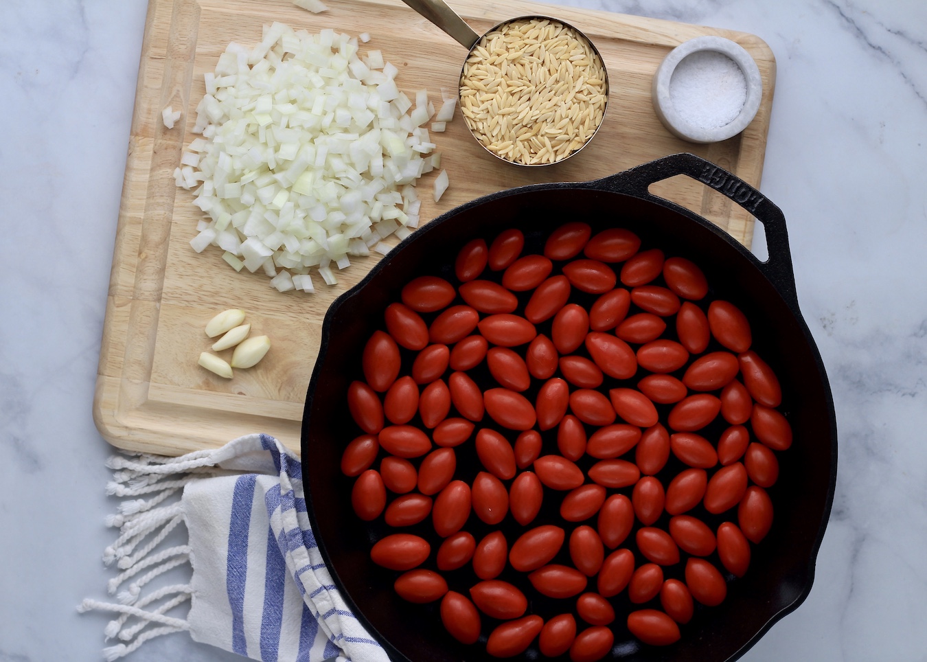 Grape tomatoes in a cast iron skillet with chopped onions, garlic, orzo, and salt on a cutting board around the skillet.