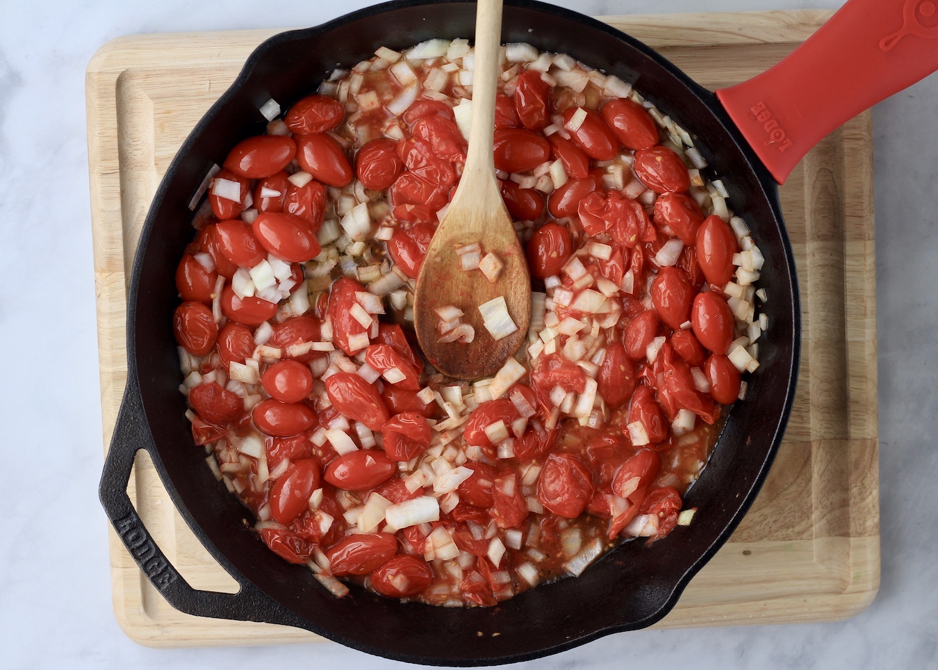 A cast iron skillet with roasted tomatoes, sauteed onion, and garlic with a wooden spoon on a wooden cutting board.