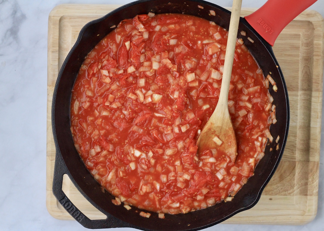 Roasted tomatoes smashed into a sauce with onion and garlic with a wooden spoon on a wooden cutting board.