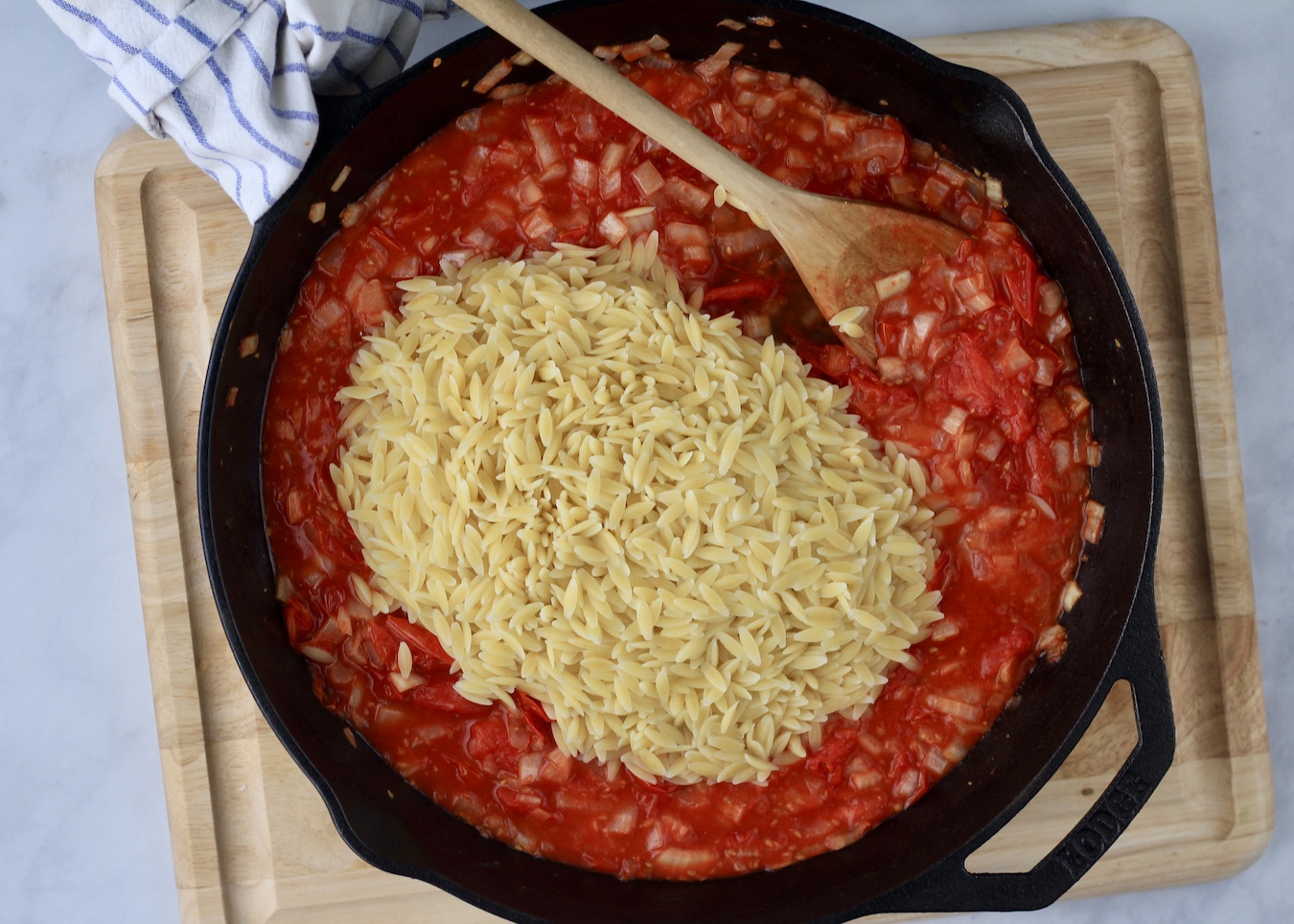 Cooked orzo added to the roasted tomato sauce with a wooden spoon on a wooden cutting board.