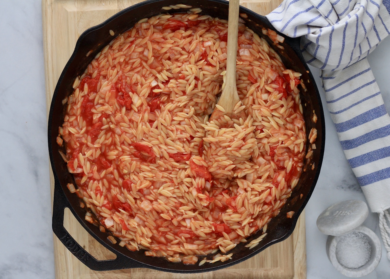 A cast iron skillet with roasted tomato orzo with a wooden spoon on a wooden cutting board next to a small pinch bowl of salt.