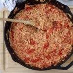 A black cast iron skillet with roasted tomato orzo and a wooden spoon on a wooden cutting board.