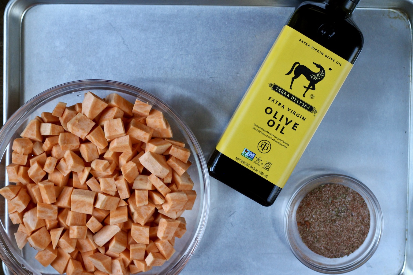 A sheet pan chopped sweet potatoes, an olive oil bottle, and a small bowl with seasoning.