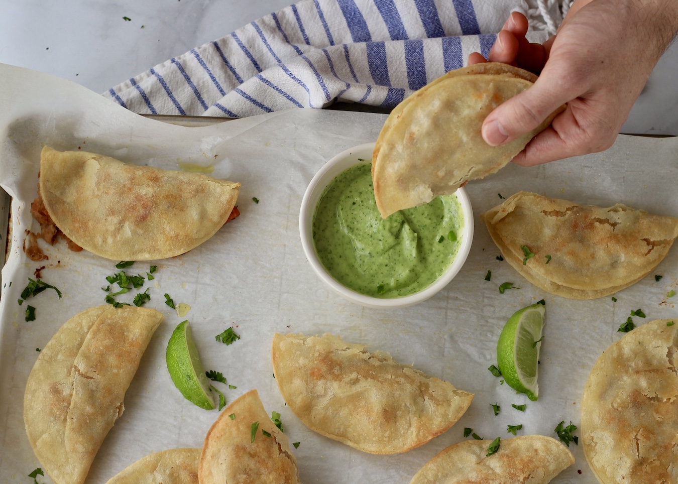A hand dipping a taco into the avocado dip with more tacos on a baking sheet in front.