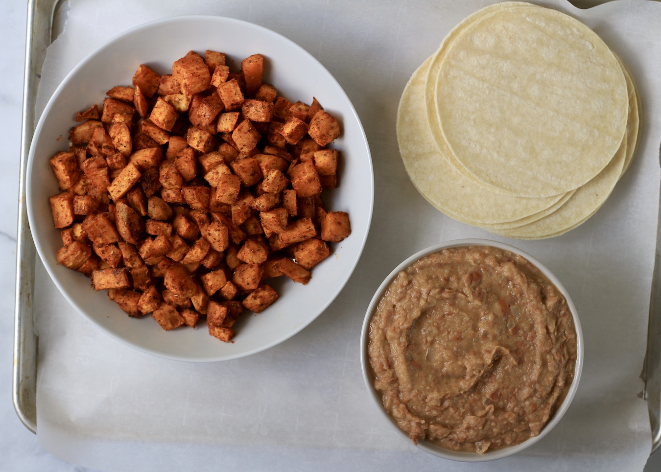 A sheet pan with parchment paper with a bowl of oven roasted sweet potatoes, a small bowl of refried beans, and a small stack of corn tortillas.