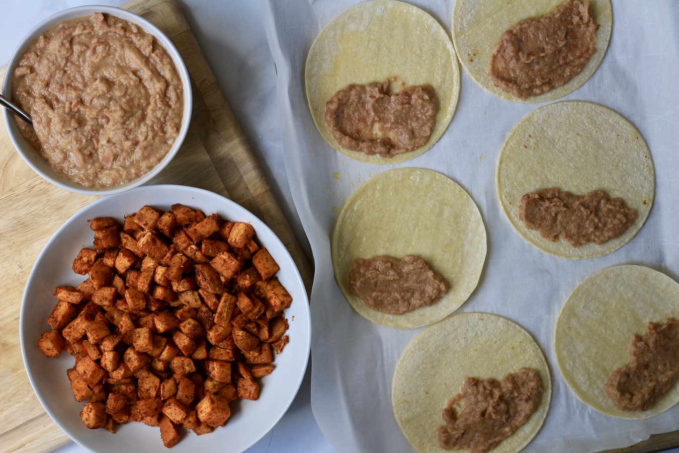 A cutting board with refried beans and sweet potatoes in bowls with a sheet pan on the right with corn tortillas with beans on one half of each corn tortilla.