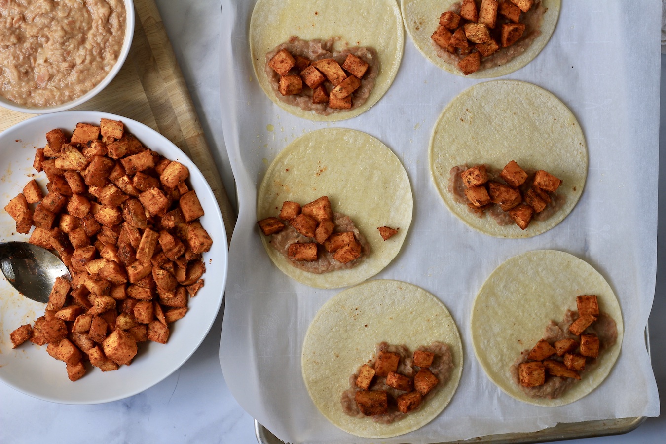 A cutting board with refried beans and sweet potatoes in bowls with a sheet pan on the right with corn tortillas with beans and potatoes.