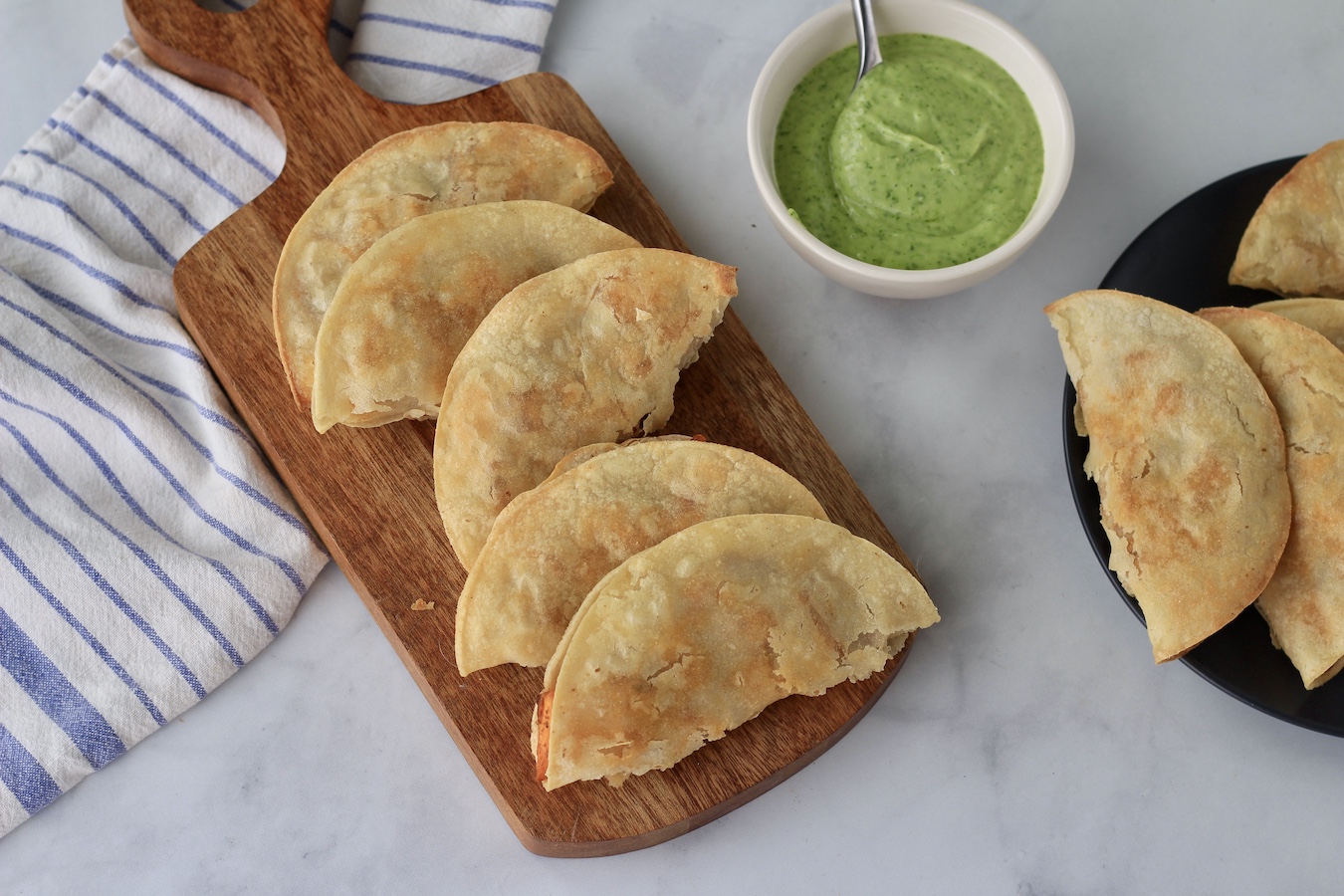 A cutting board on the left with sweet potato tacos, a bowl of avocado dressing in the middle, and a small plate of sweet potato tacos on the right.