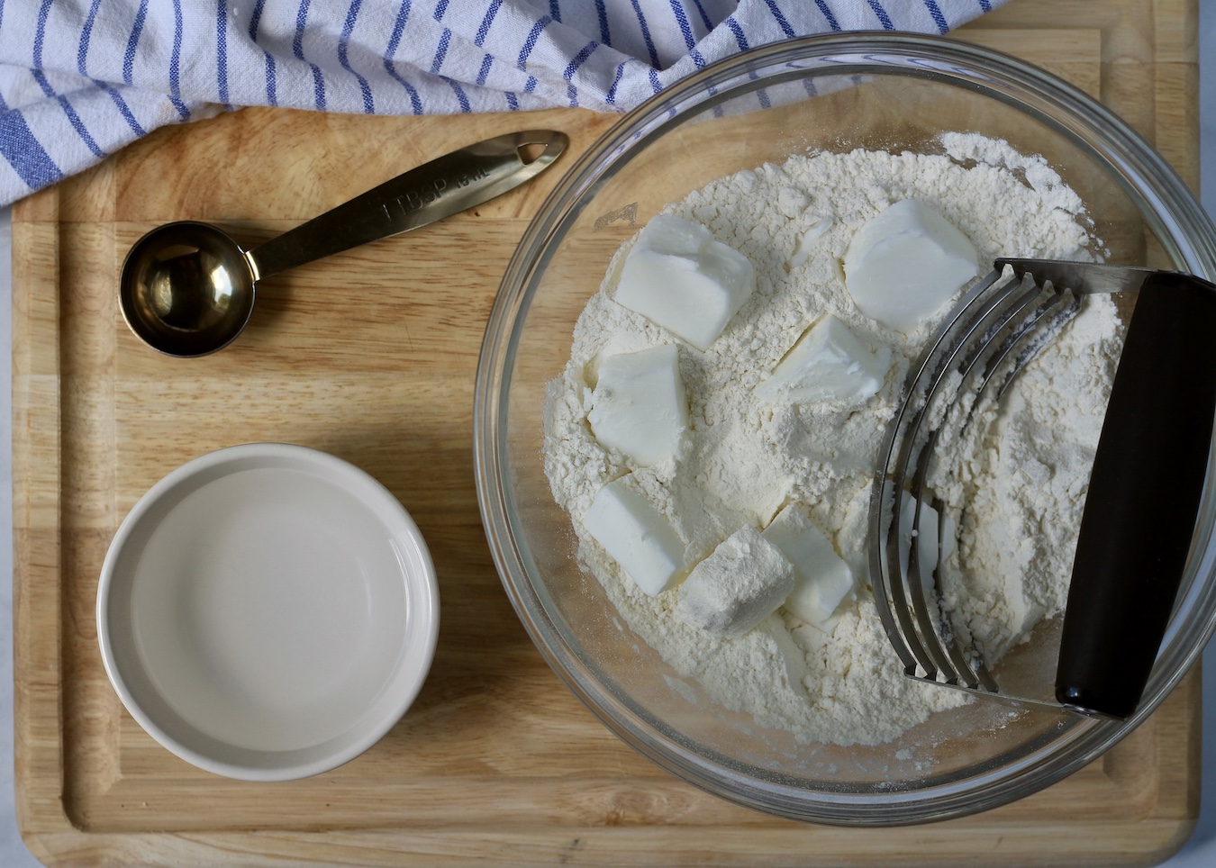 A large bowl with flour and shortening on a cutting board with ice water and a tablespoon.