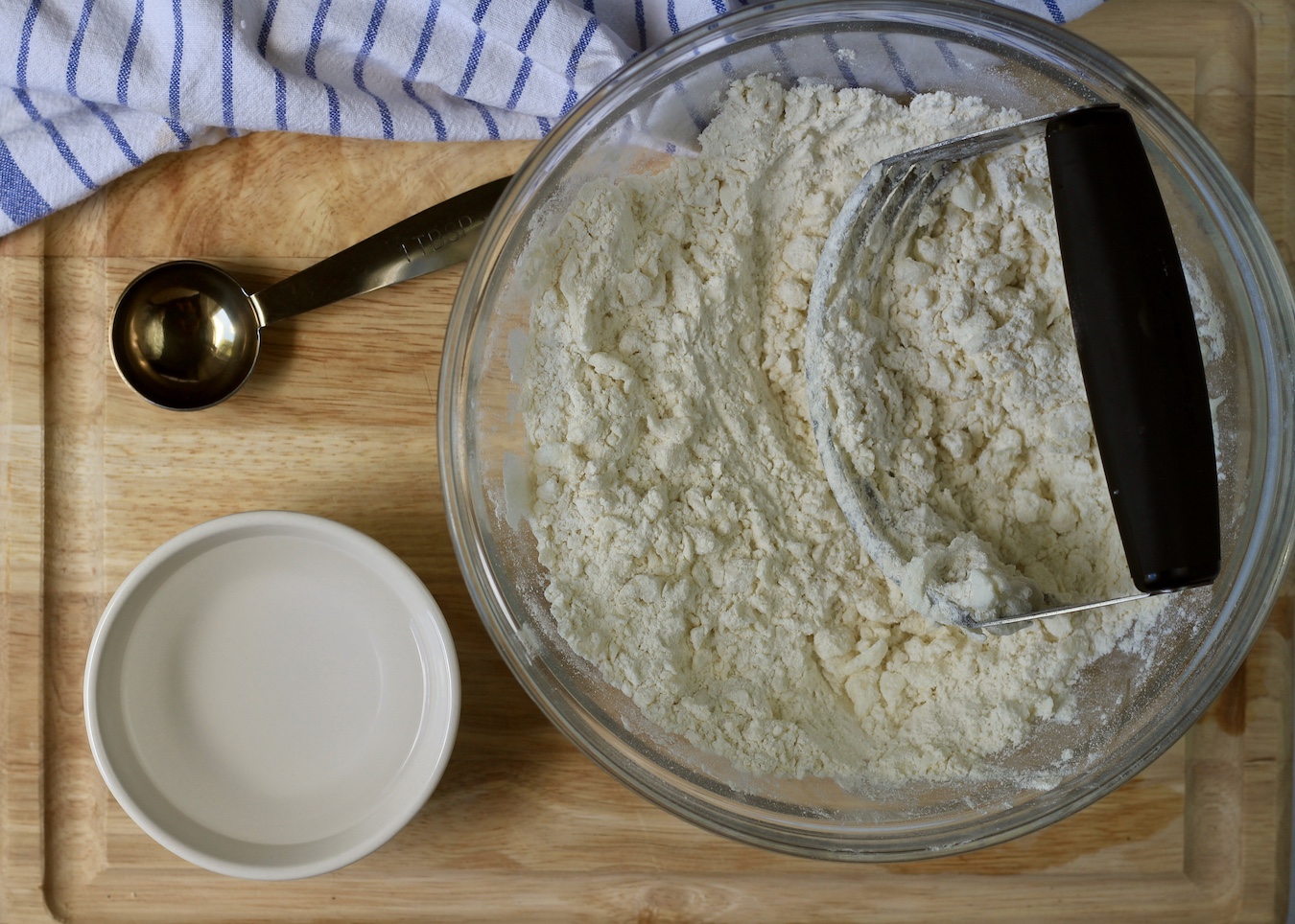 A large bowl with shortening cut into flour with a pastry blender on a wooden cutting board with a bowl of ice water and a tablespoon.