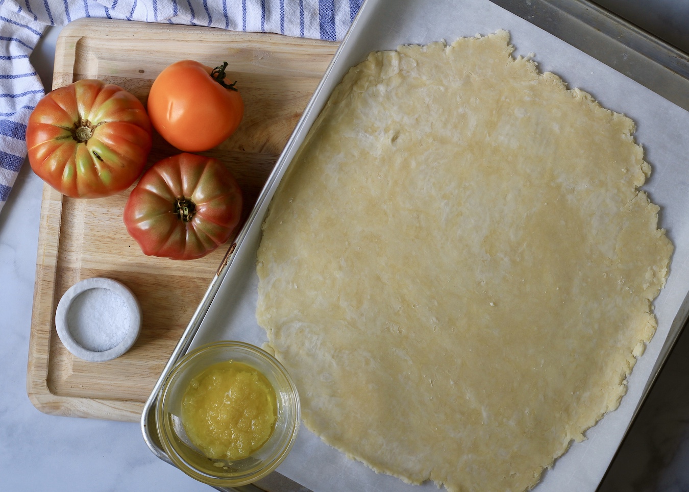 A large sheet pan with rolled pastry dough on parchment paper, a small bowl of garlic and olive oil, three heirloom tomatoes to the left and a small bowl of salt to the bottom left.