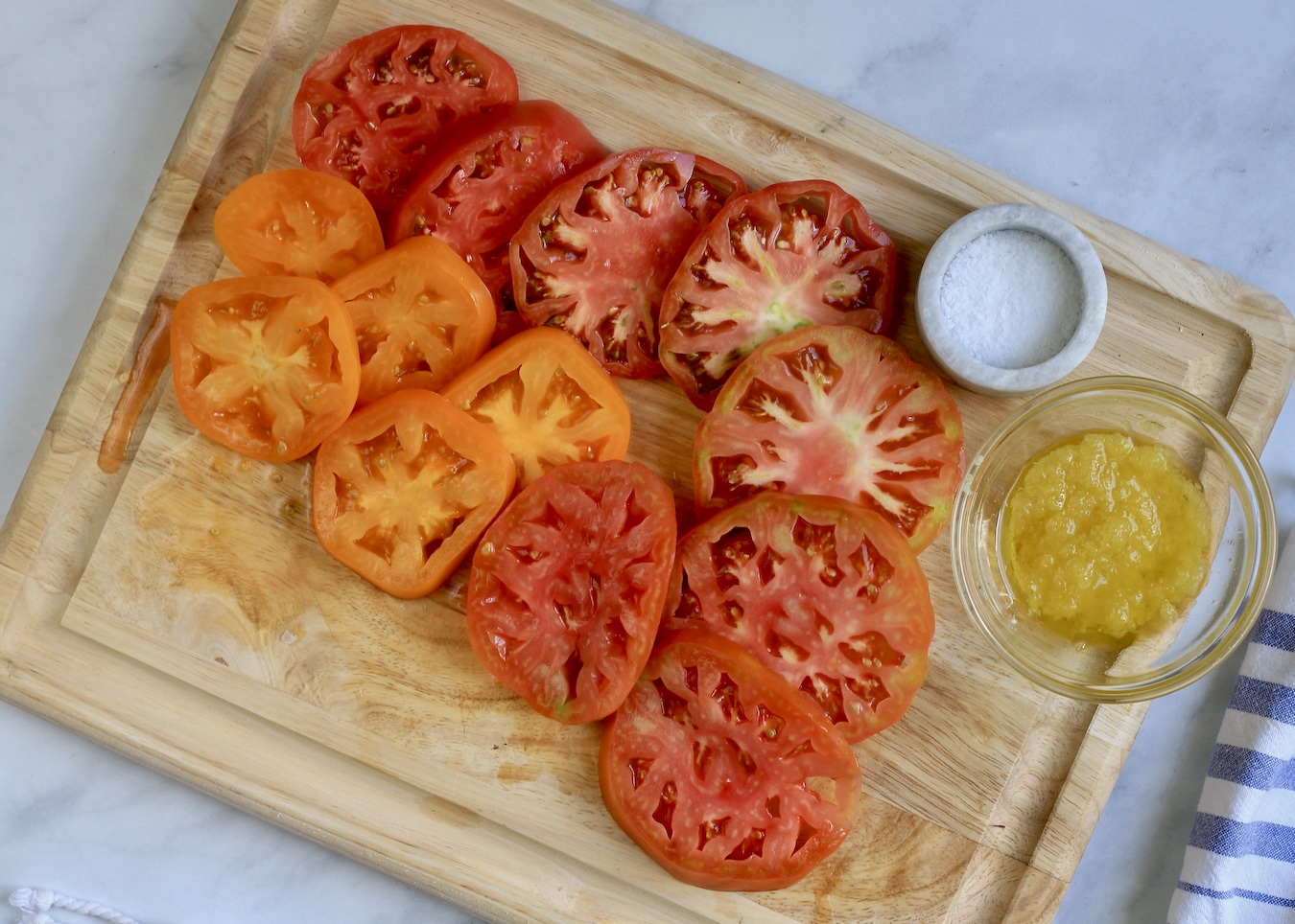 A wooden cutting board with sliced heirloom tomatoes and a small bowl of garlic with olive oil and a pinch bowl with salt.