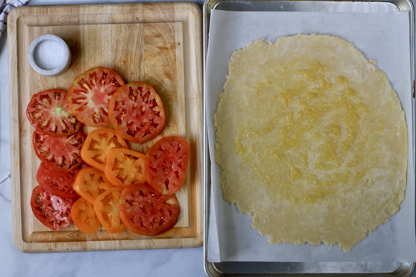 A cutting board with sliced tomatoes and a salt bowl on the left with a rolled out pastry on the right.