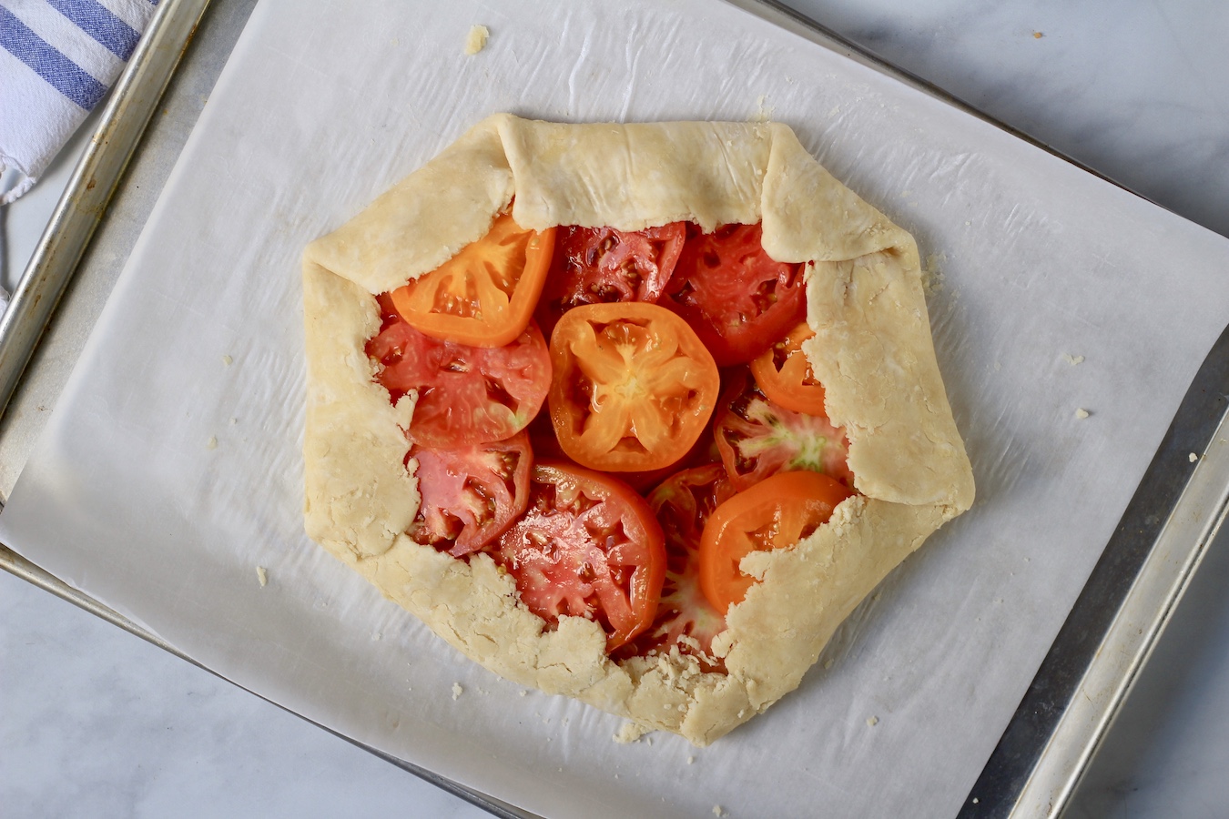 The edges of the galette being folded over the edges of the circle of tomatoes on a parchment paper on a baking sheet.