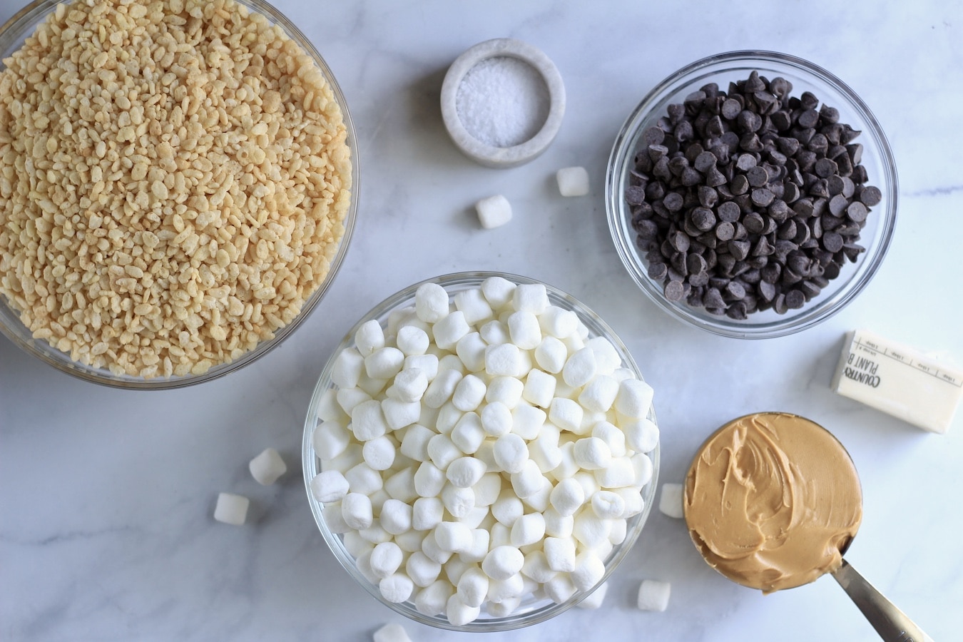 From left to right a glass bowl with rice krispie cereal, a glass bowl with marshmallows, a stone bowl with salt, a glass bowl with chocolate chips, 1 cup measuring cup with peanut butter and a half stick of vegan butter.