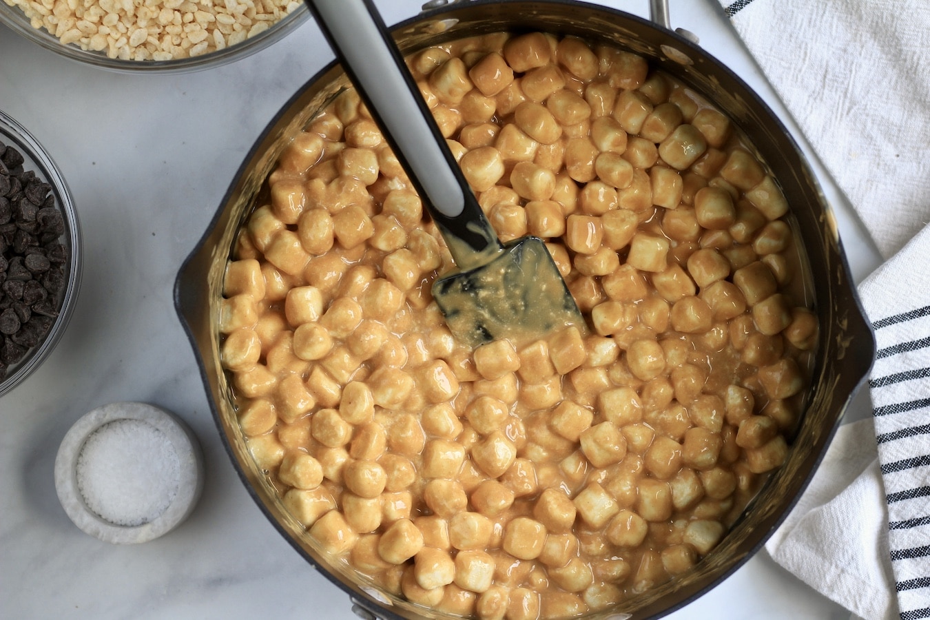 A non-stick pot with melted peanut butter and marshmallows with a black rubber spatula next to a stone bowl with salt.