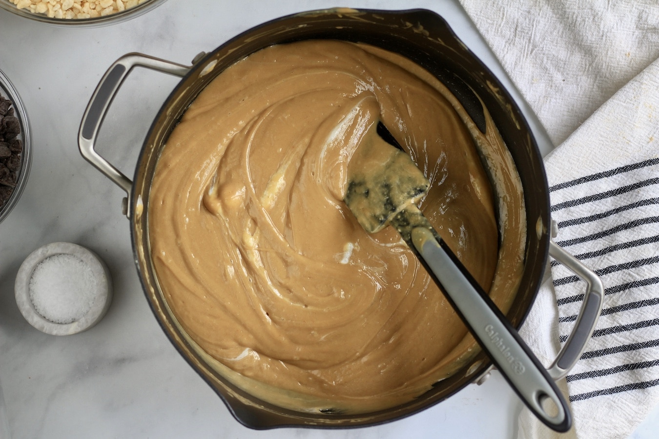 A non-stick pan with melted peanut butter and marshmallows, with a black rubber spatula next to a stone bowl with salt.