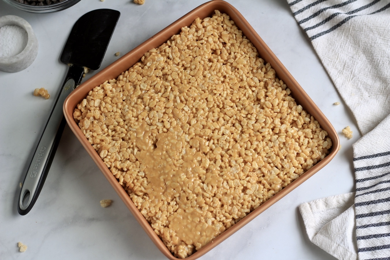 Peanut butter rice krispies pressed into a copper pan with a black rubber spatula to the left.