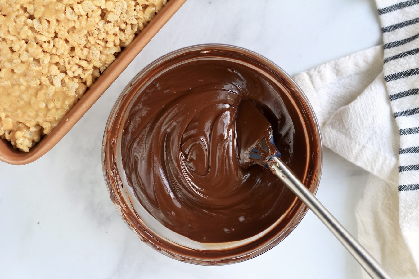 A small glass bowl with melted chocolate and a small rubber spatula next to the peanut butter rice krispies.