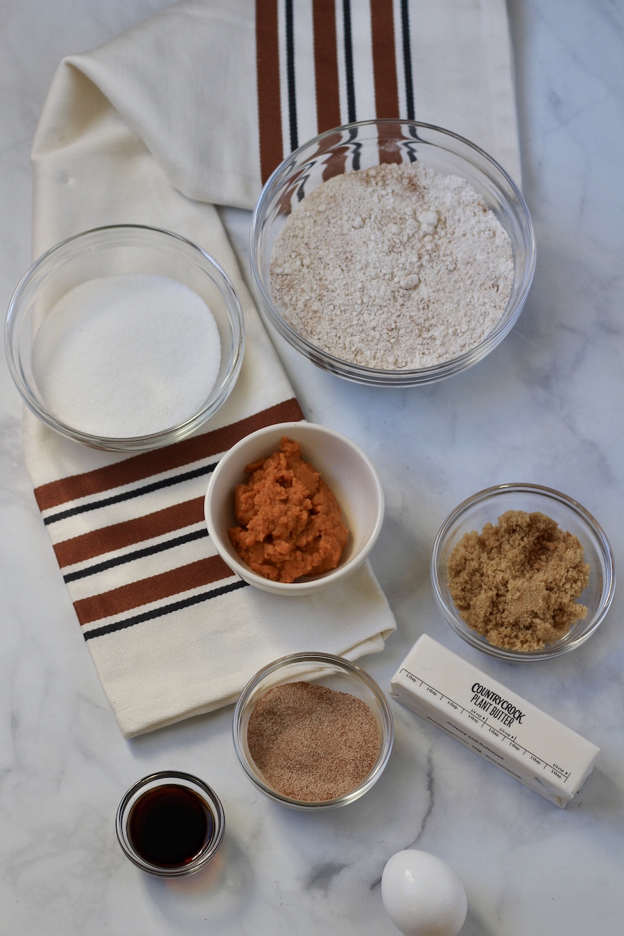 Glass bowls with the ingredients for pumpkin spice snickerdoodle cookies on a marble counter with an orange and brown striped kitchen towel.