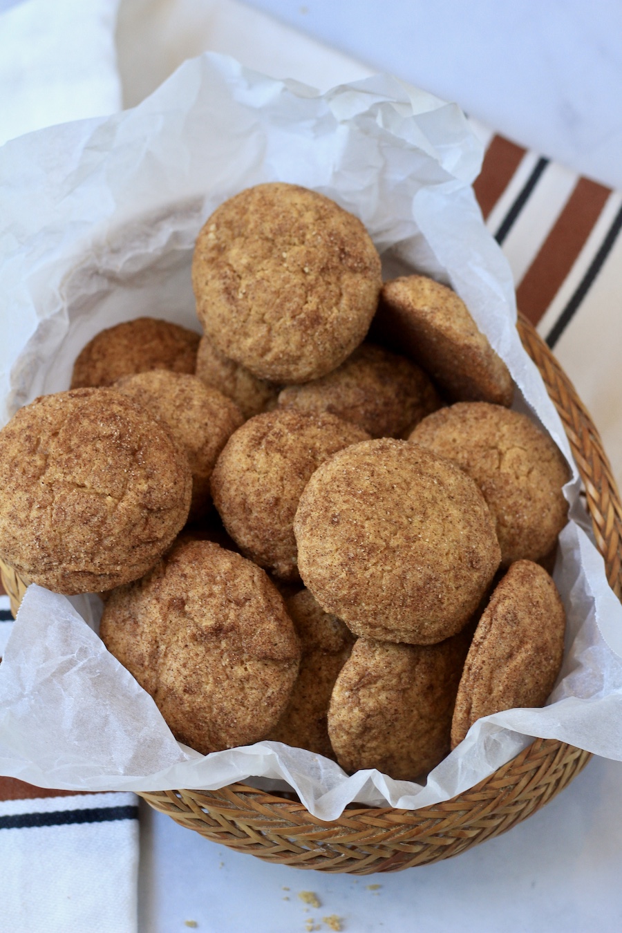 A woven basket with parchment paper filled with pumpkin spice snickerdoodles on a marble counter with a brown and cream dish towel.