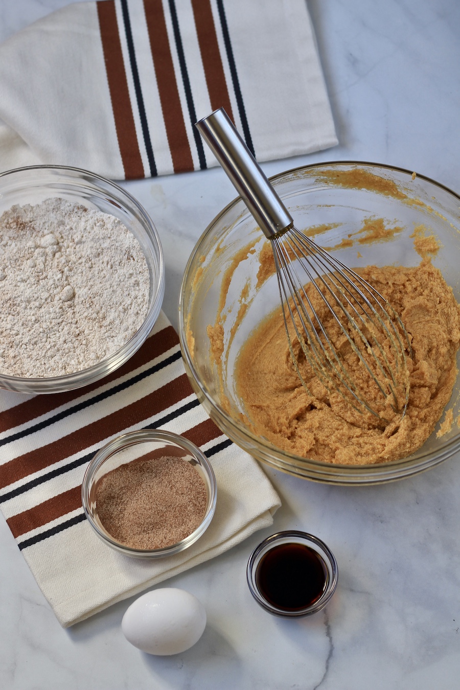 A pumpkin sugar mixture in a glass bowl with the flour mixture to the back and a small bowl with vanilla and an egg in the front on a marble counter.