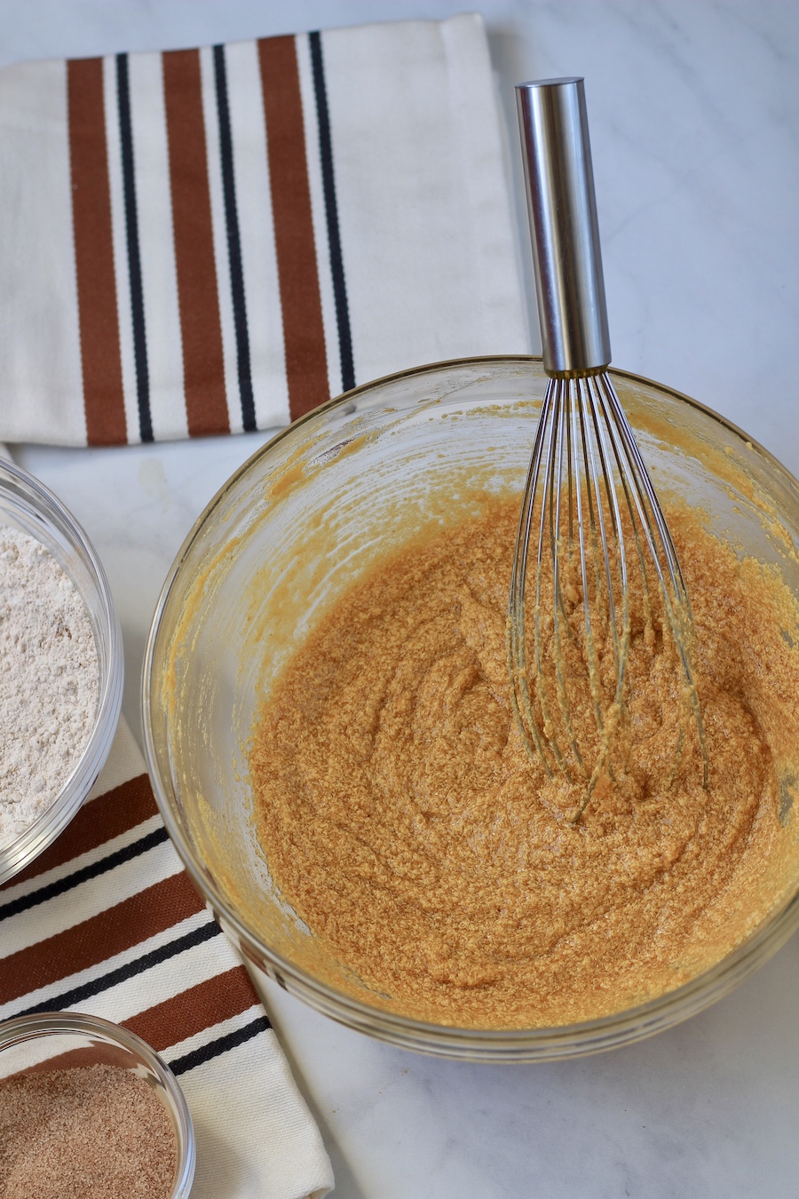 The wet ingredients of the pumpkin spice snickerdoodles in a glass bowl with a whisk on a marble counter.