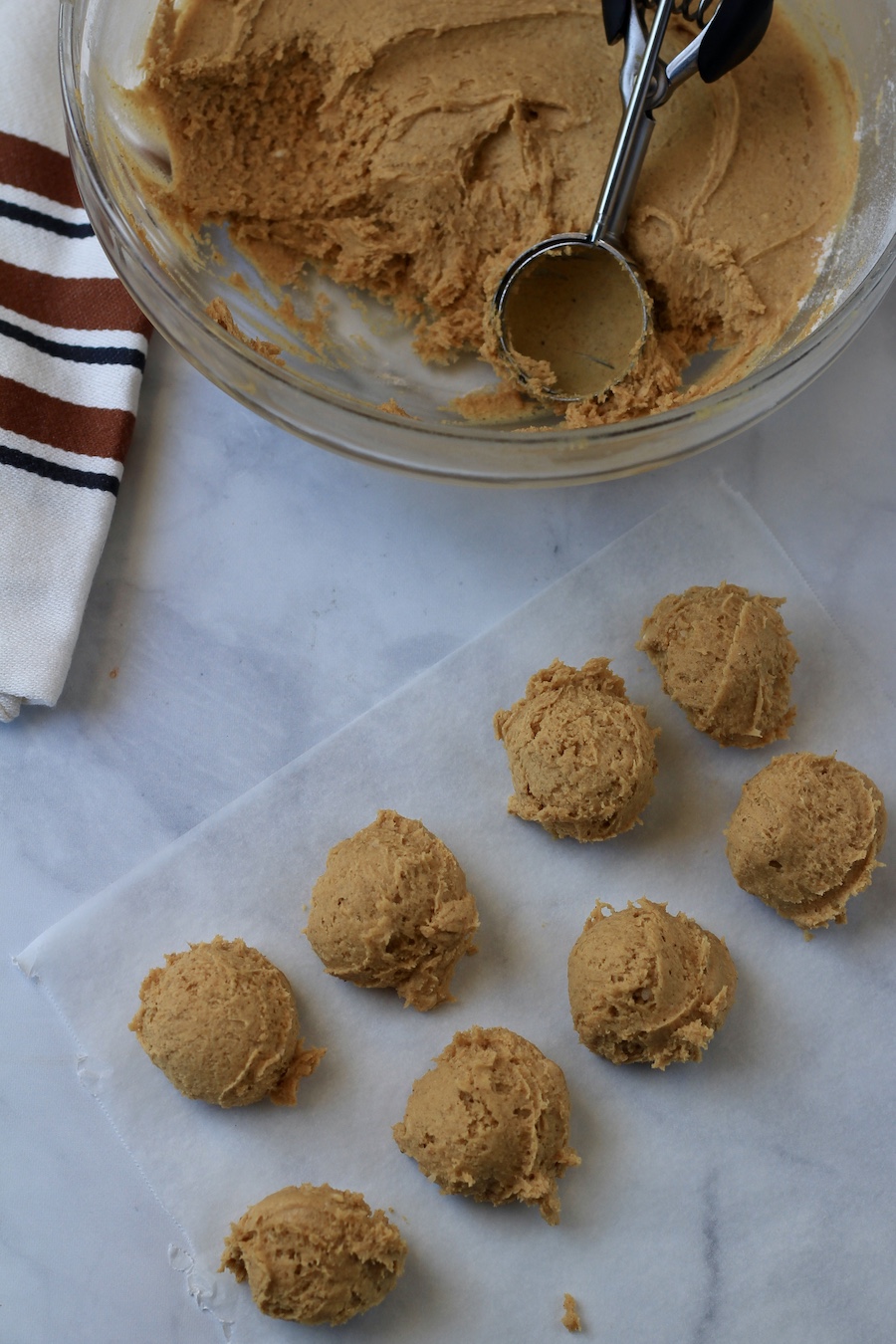 Pumpkin spice snickerdoodle cookie dough in a bowl with a cookie scoop and scoops on a parchment paper.