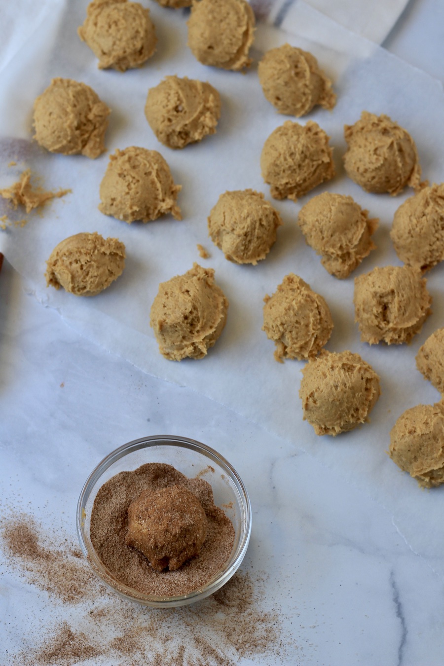 Scoops of pumpkin spice snickerdoodle cookie dough on parchment paper with a small bowl of cinnamon sugar coating.