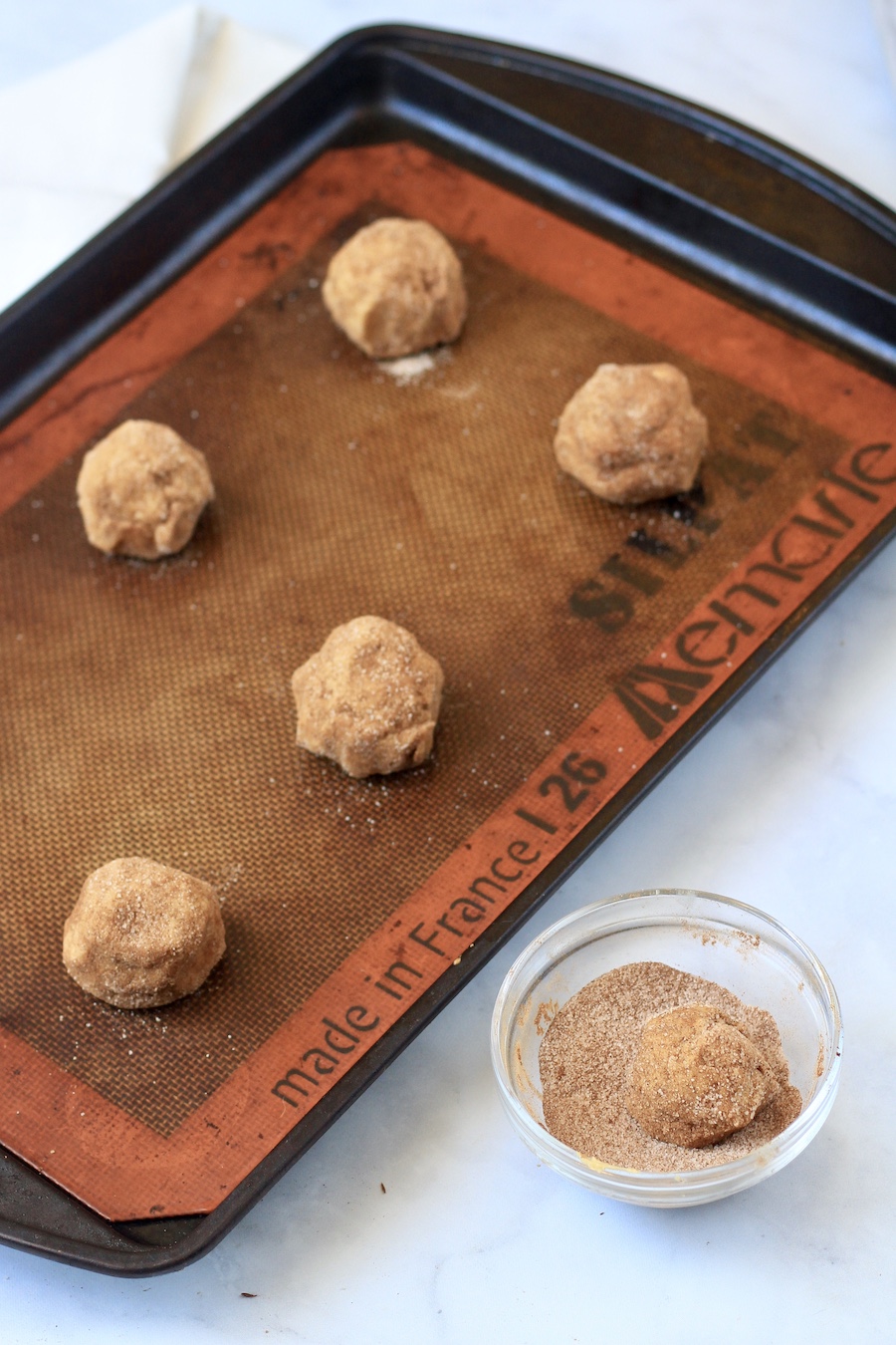 A cookie sheet lined with a silicone mat with cookie dough balls rolled in cinnamon sugar with a small bowl of cinnamon sugar in the front.