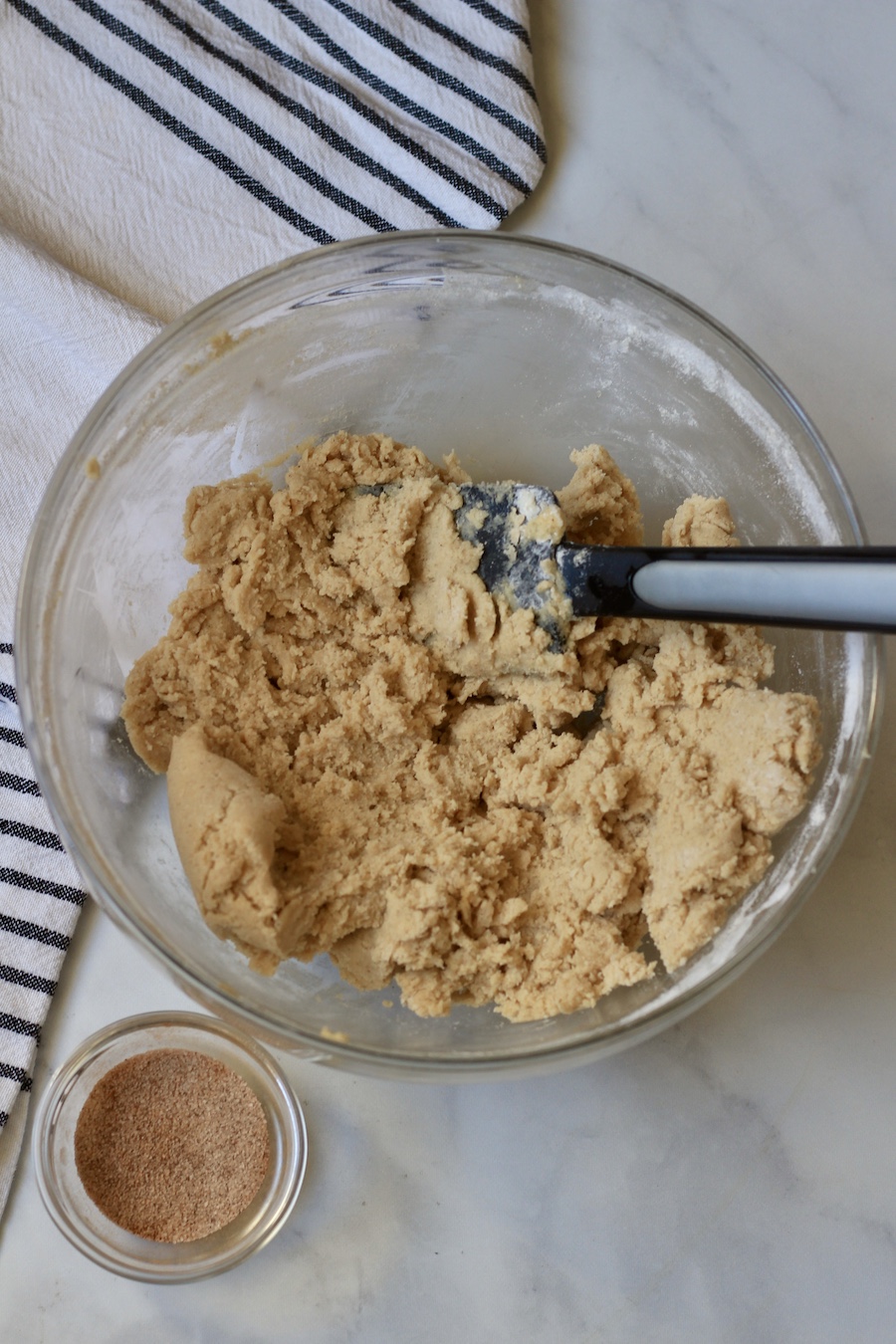 A glass bowl with snickerdoodle cookie dough and a rubber spatula with a small bowl of cinnamon sugar to the bottom left.
