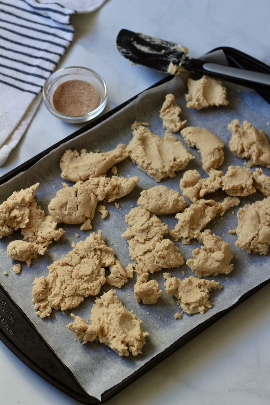 Drops of the snickerdoodle cookie dough on a parchment paper lined cookie sheet with cinnamon sugar in a small bowl to the left.